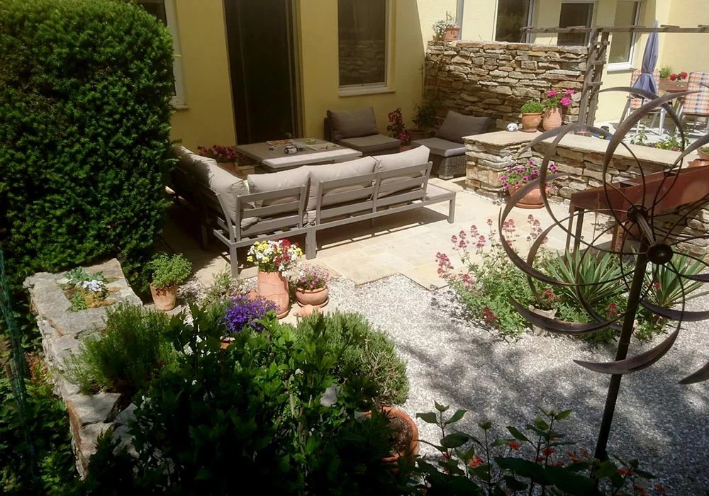 Sehr schöne Terrasse mit gemütlichen Gartenmöbel mit Reihenhaus im Hintergrund. Die Terrasse ist liebevoll dekoriert, man sieht viele bepflanzte Terracotta-Töpfe und eine filigrane Skulptur aus Metall.