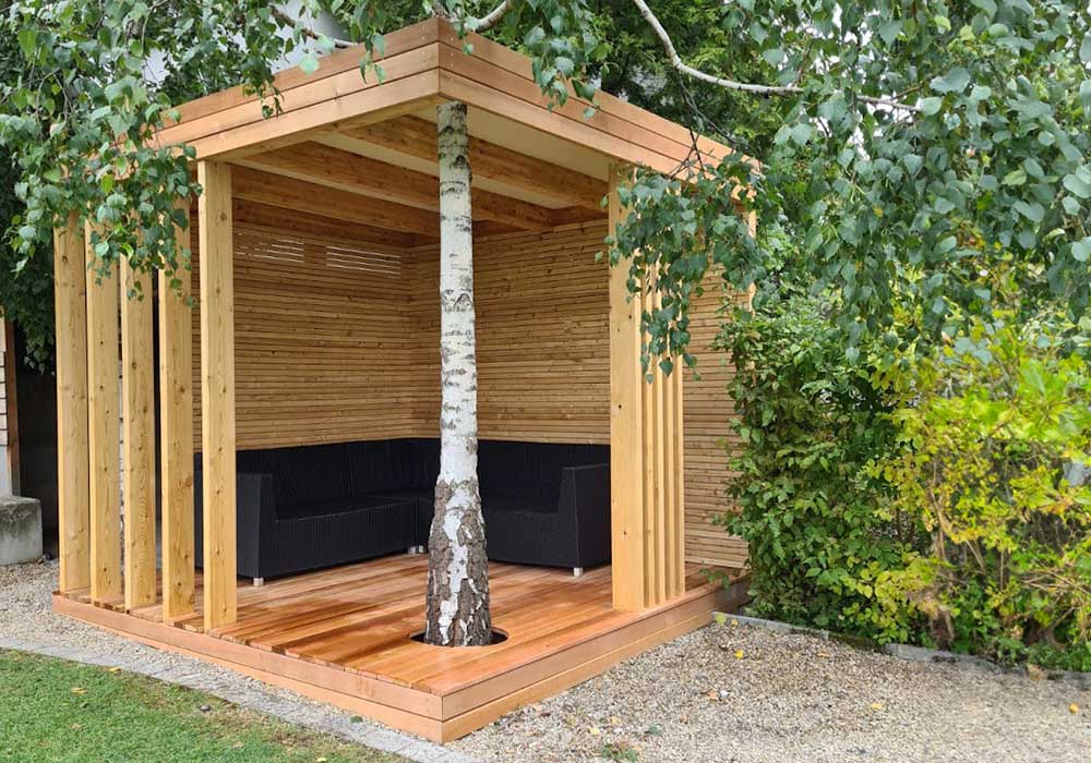 Loungebereich aus Holz gestaltet. Mit integriertem Baum, der vorne zu sehen ist. Im Loungebereich stehen große Gartenmöbel.