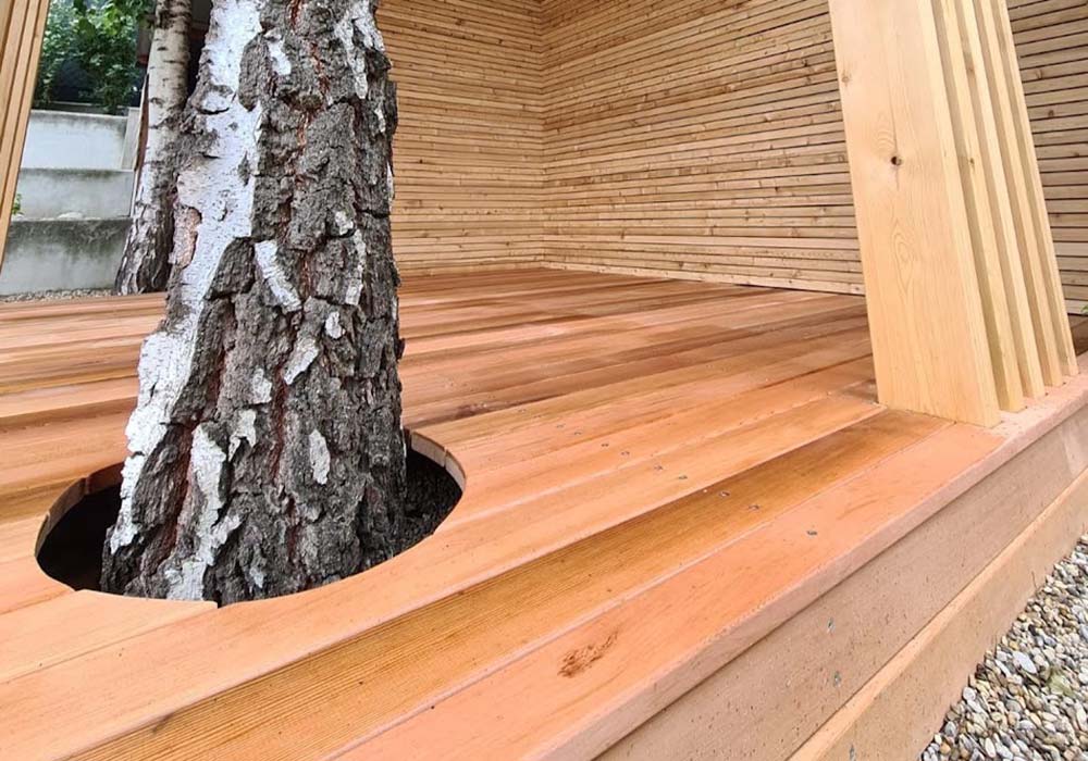 Detailansicht von einem Loungebereich der komplett aus Holz gebaut wurde. Im Bau wurde ein Baum integriert, er wächst durch einen Ausschnitt im Boden.