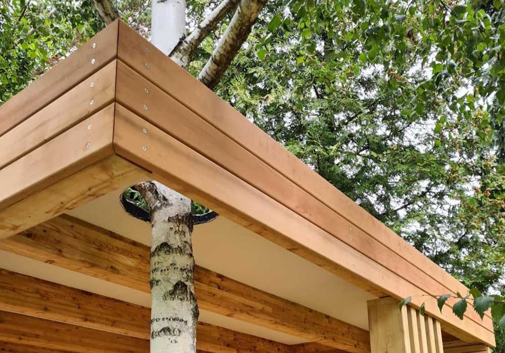 Detailansicht von einem Loungebereich der komplett aus Holz gebaut wurde. Im Bau wurde ein Baum integriert, er wächst durch einen Ausschnitt im Dach.