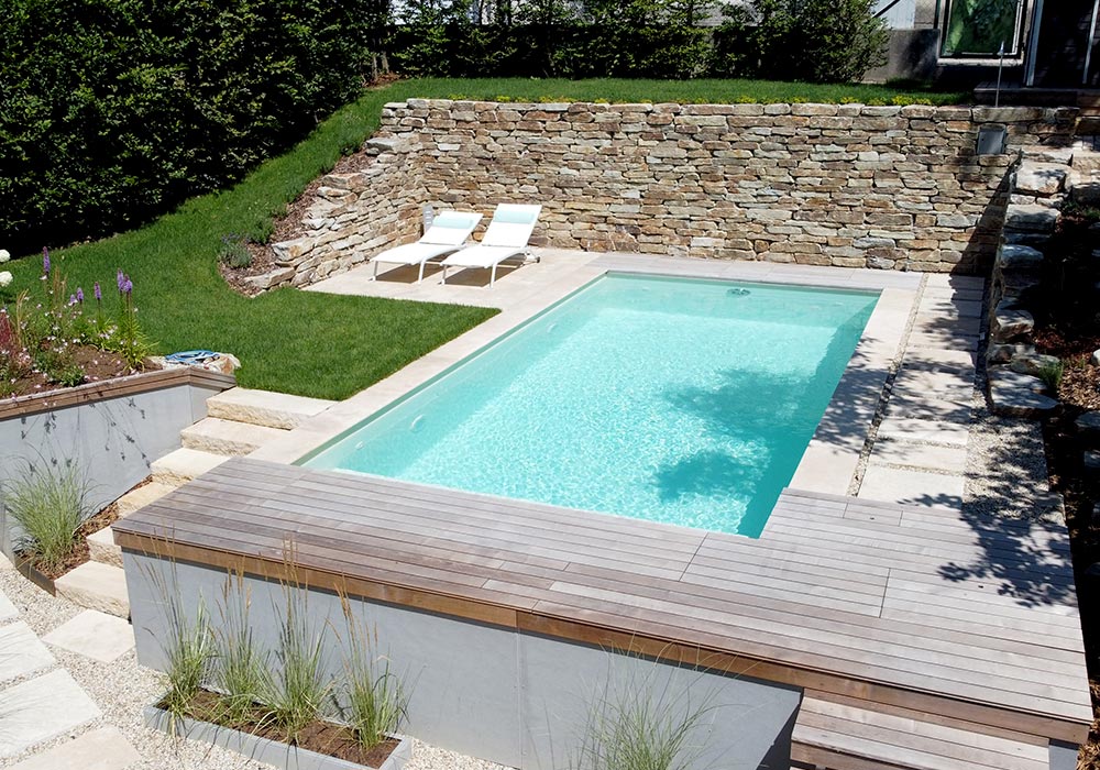 Ein schöner sonniger Garten, mit Pool eingerahmt von einer Holz- und Steinterrasse. Hinten gibt es eine Steinmauer, links stehen zwei Sonnenliegen