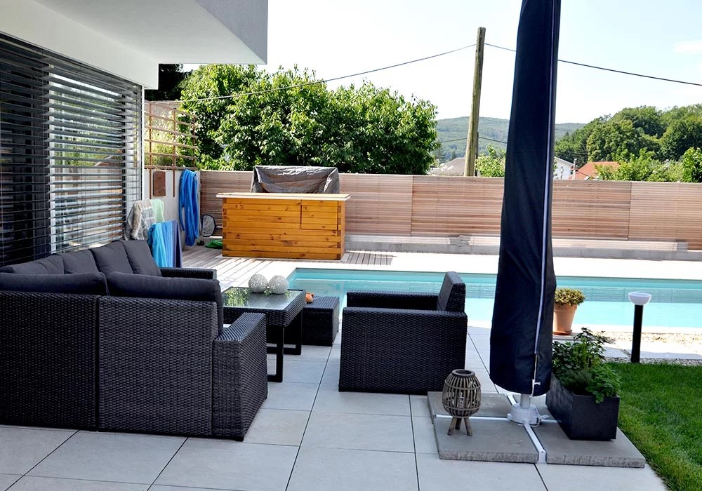 Detailansicht Garten mit großer Terrasse. Auf der Terrasse stehen ein gemütliche Gartenmöbel und ein abgedeckter Sonneschirm. Im Hintergrund sieht man einen Pool der mit einer Sichtschutzwand aus Holz nach außen geschützt ist.