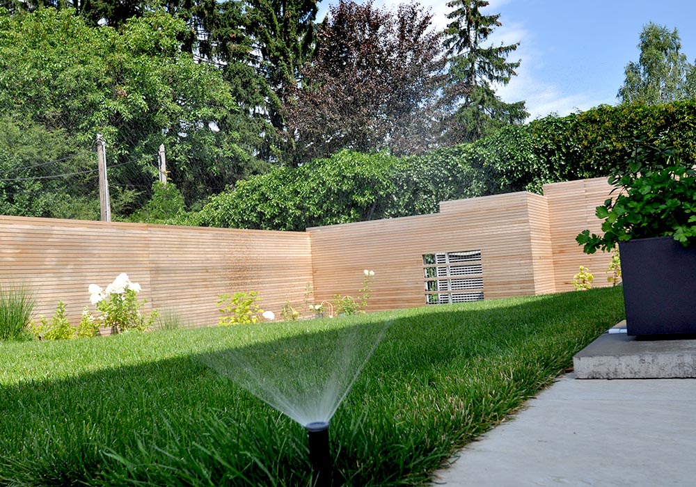 Detailansicht eines Gartens mit Sichtschutzwand aus Holz im Hintergrund. Vorne sieht man ganz nah einen Gartensprinkler.